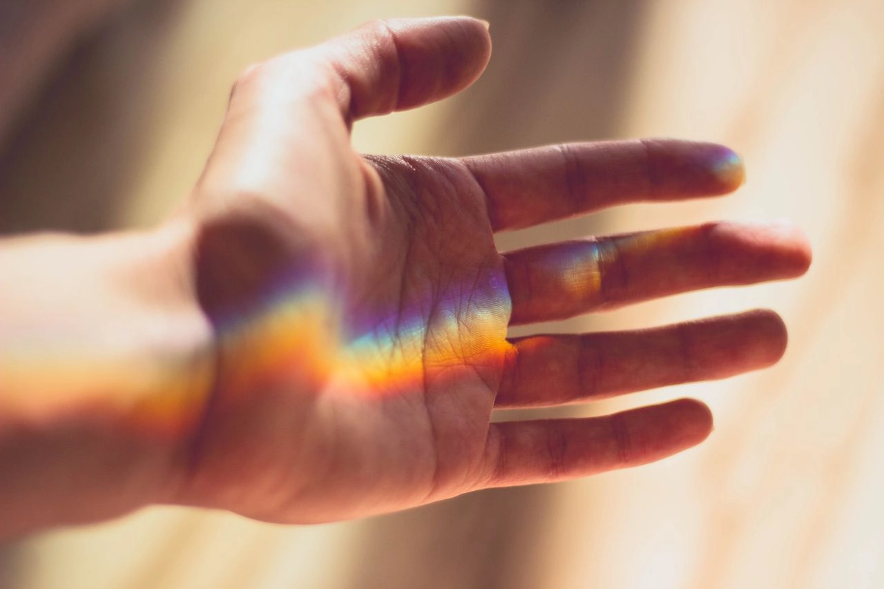 een hand in de zon, waar door reflectie een regenboog verschijnt op de palm van de hand